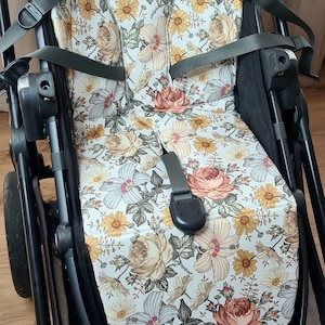 Könnte beinhalten: Ein schwarzer Kinderwagen mit einer Sitzbezug mit Blumenmuster. Der Bezug hat einen weißen Hintergrund mit verschiedenen Blumen in den Farben Rosa, Gelb und Braun.