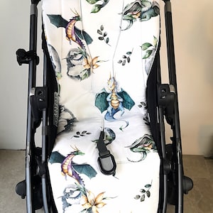 Könnte beinhalten: Ein weißer Kinderwagen-Sitzbezug mit einem Aquarell-Drachen-Druck. Der Bezug zeigt mehrere Drachen in Blautönen, Grün- und Violetttönen, mit gelben Blumen und grünen Blättern.