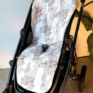 Könnte beinhalten: Ein schwarzer Kinderwagen mit einer weißen und braunen gemusterten Sitzbezug. Der Bezug hat ein geometrisches Muster aus Kreisen und Linien.