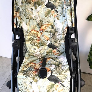 Könnte beinhalten: Ein schwarzer Kinderwagen mit einem tropischen bedruckten Verdeck und Sitzbezug. Der Druck zeigt Tukane, Papageien und Palmenblätter auf einem hellgrünen Hintergrund.