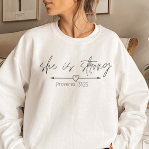 以下が含まれることがあります： 「she is strong」というテキストと、右を向いた矢印のある小さなハートが書かれた白いスウェットシャツ。テキストの下には「Proverbs 3125」とあります。