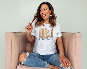 May include: Light gray t-shirt with the word "BOOKTROVERT" printed below a stack of illustrated books with floral accents. The books are in various colors, including pink, green, and orange. The shirt is short-sleeved.