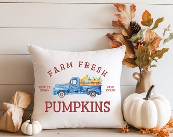 May include: White throw pillow with a blue pickup truck carrying pumpkins. The truck has the words "Farm Fresh" above it and "Locally Grown" and "Hand Picked" on either side. The words "Pumpkins" are below the truck.