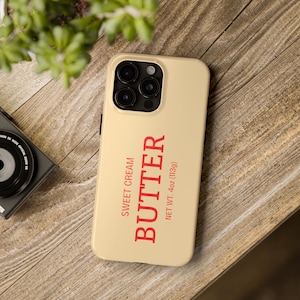 May include: A beige phone case with a red "BUTTER" label, featuring "SWEET CREAM" and "NET WT. 4oz (113g)" text. The case has a camera module with multiple lenses. The phone case is on a wooden surface.