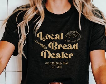 May include: A black t-shirt with the text "Local Bread Dealer" in gold lettering. The text is in a retro font style. There is a drawing of a loaf of bread and a whisk next to the text. The text "Custom Bakery Name Est. 2025" is below the main text.