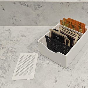May include: White plastic tea bag organizer with a black grid insert. The organizer is filled with tea bags, some with floral patterns and some with the word "ETOO" printed on them.