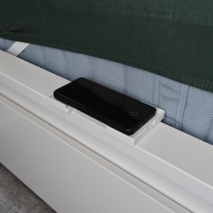 May include: A white phone holder attached to the side of a bed frame. A black smartphone is resting in the holder.