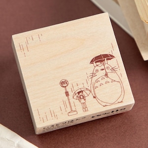 May include: A wooden stamp featuring a brown illustration of a large, round creature with an umbrella, a small person with an umbrella, and a bus stop in the rain. The stamp has Japanese text at the bottom.