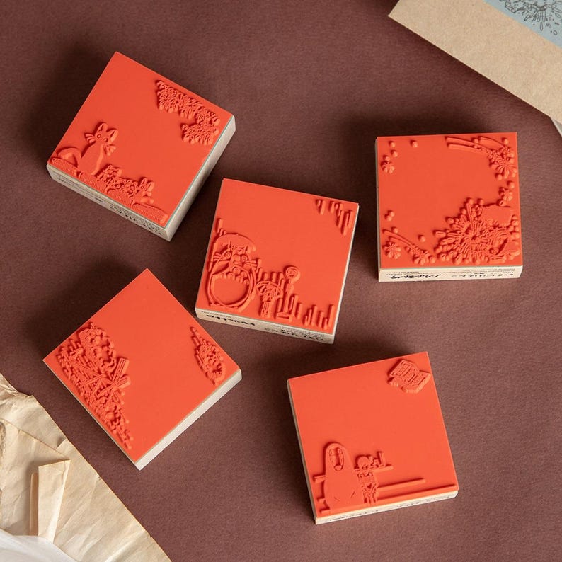 May include: Five square, red rubber stamps with various designs, including floral, architectural, and whimsical illustrations. The stamps are mounted on wooden blocks, and are arranged on a brown surface.