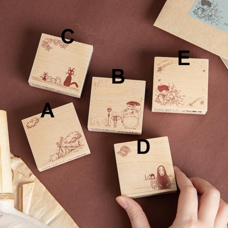 May include: A collection of five wooden stamps with various illustrations. The stamps feature designs of a cat, a character with an umbrella, a girl with flowers, and other whimsical scenes. The stamps are square and made of light-colored wood.