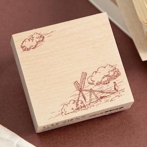 May include: A wooden stamp with a brown illustration of clouds, a windmill, and a person. The stamp has a light wood grain and a rectangular shape. The illustration is detailed and appears to be hand-drawn.