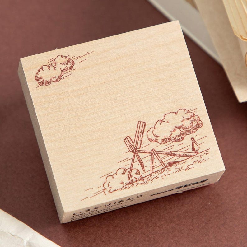 May include: A wooden stamp with a brown illustration of clouds, a windmill, and a person. The stamp has a light wood grain and a rectangular shape. The illustration is detailed and appears to be hand-drawn.