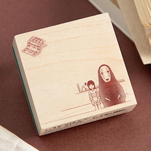 May include: A wooden stamp featuring a brown illustration of characters from a Japanese animated film. The stamp includes text and a detailed drawing of a girl and a tall, faceless figure. The stamp has a green rubber base.