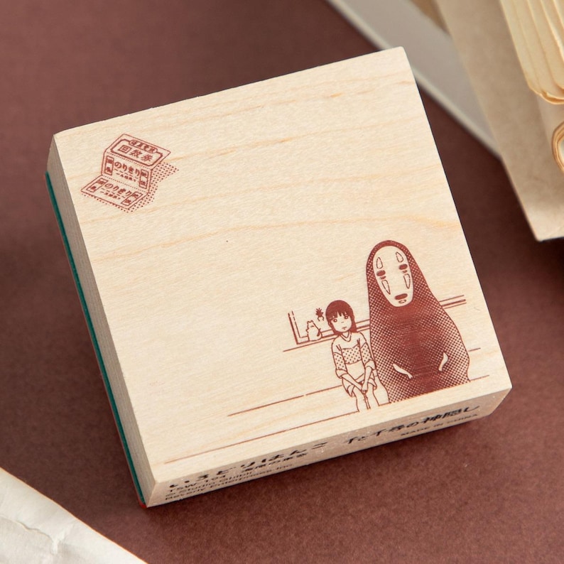 May include: A wooden stamp featuring a brown illustration of characters from a Japanese animated film. The stamp includes text and a detailed drawing of a girl and a tall, faceless figure. The stamp has a green rubber base.