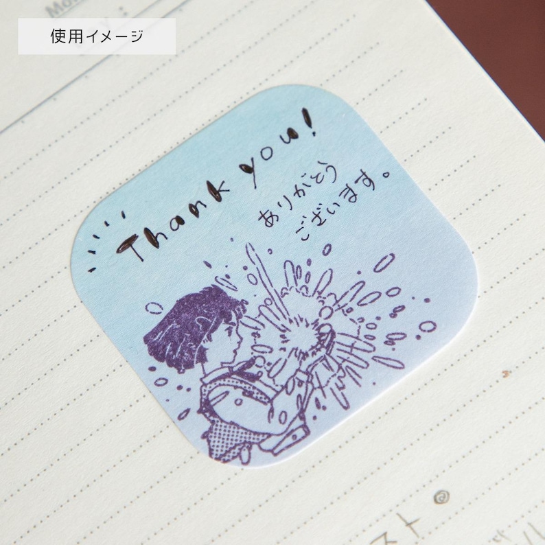 May include: A light blue sticker with rounded corners featuring a purple illustration of a person and the words "Thank you!" in English and Japanese. The sticker has a hand-drawn style and is placed on a lined notebook page.