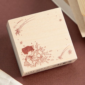May include: A wooden stamp featuring a whimsical illustration of a person interacting with a burst of light and shooting stars. The design is in reddish-brown ink on a light wood block. The stamp has Japanese text along the bottom edge.