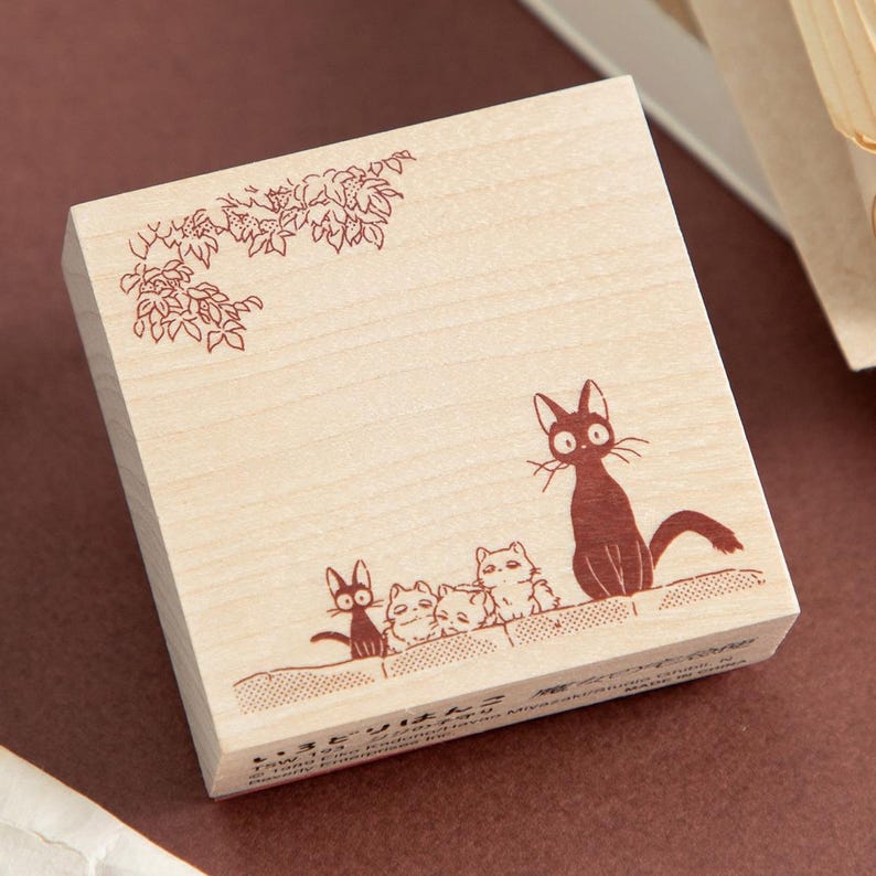 May include: Wooden stamp featuring a brown illustration of several cats and floral elements. The cats are depicted in a row, with one cat standing out. The stamp has a light wood finish.