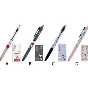 May include: Four different pens featuring Peanuts characters. The pens are beige, grey, black, and pink. The pens have the text "デザイン" on them. The pens are next to small cards with Peanuts characters on them.