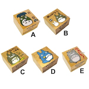 Peut inclure: Cinq tampons en caoutchouc carrés en bois étiquetés de A à E, chacun présentant une illustration différente de personnages Totoro avec du texte japonais. Les tampons ont des bases en bois brun clair et montrent diverses scènes avec Totoro, un parapluie, une grenouille et d'autres petits esprits de la forêt en encre noire, blanche, bleue et verte.