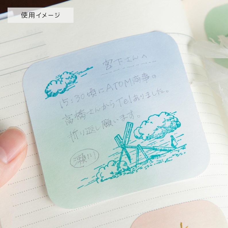 May include: A square, gradient-colored sticky note with handwritten Japanese text and illustrations in teal ink. The note includes a drawing of a landscape with clouds and a seesaw. The text is in Japanese.