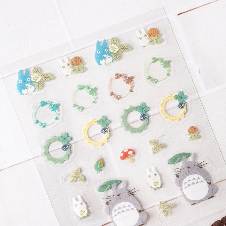Peut inclure: Une feuille d'autocollants d&eacute;coratifs avec des personnages de dessins anim&eacute;s et des motifs de couronnes florales. Les autocollants comprennent des personnages bleus et gris, des feuilles vertes et un champignon rouge. Les autocollants sont sur un fond transparent.