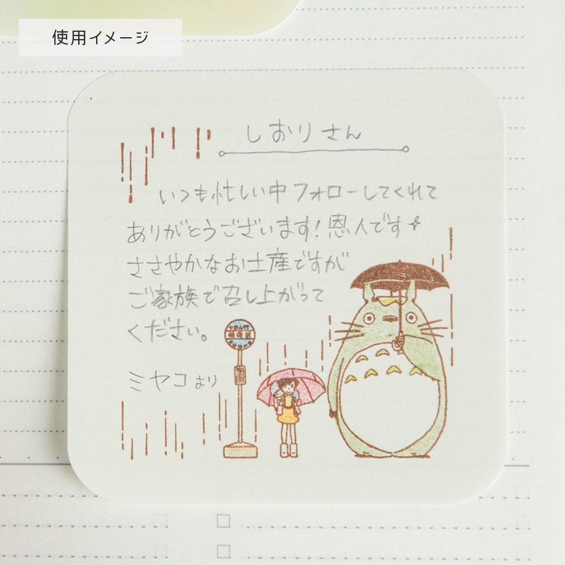 May include: A square, off-white sticky note with Japanese text and a drawing of Totoro and a girl under an umbrella in the rain. The note also features a bus stop and vertical lines.