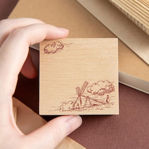 May include: A square wooden stamp with a brown illustration of a windmill, clouds, and a person. The stamp is held by a hand, with a neutral background of books and paper.