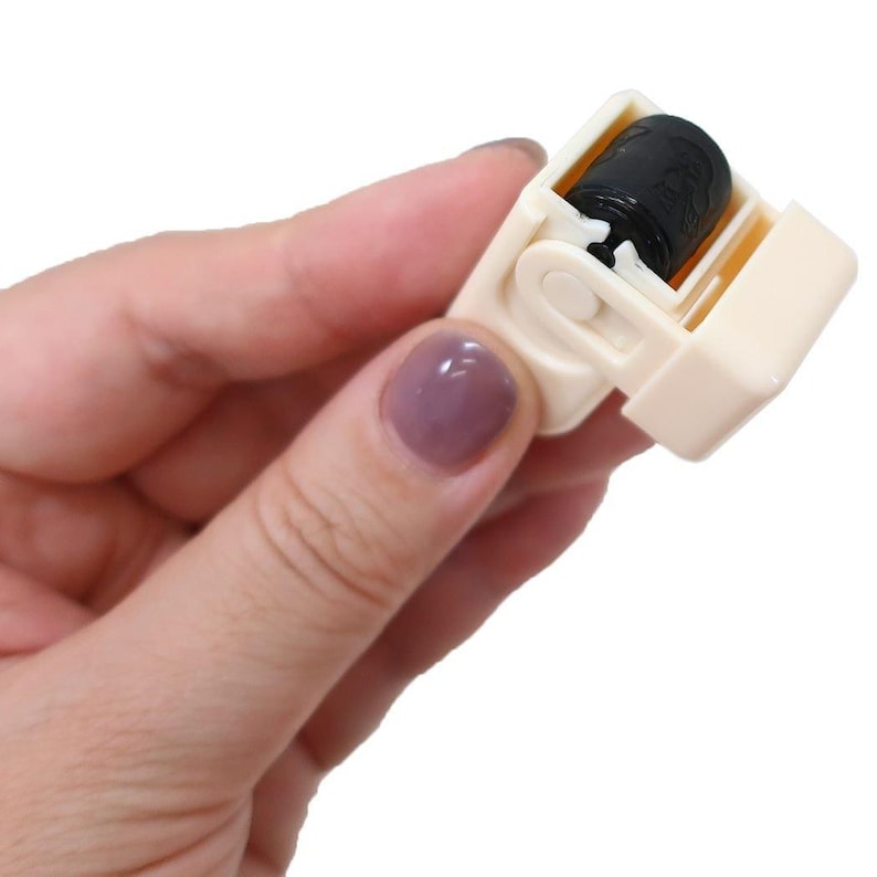 Puede incluir: Un peque&ntilde;o rodillo de pl&aacute;stico blanquecino con una superficie adhesiva negra y cil&iacute;ndrica. El rodillo se mantiene abierto, revelando la superficie pegajosa, y est&aacute; dise&ntilde;ado para eliminar pelusas y residuos de la tela.