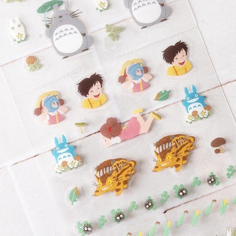 Peut inclure: Feuille d'autocollants transparents avec divers personnages de dessins anim&eacute;s, dont une grande cr&eacute;ature grise avec un parapluie en feuille, une cr&eacute;ature bleue et des enfants. Les autocollants sont color&eacute;s et ont un design fantaisiste, avec des accents floraux et de feuilles.