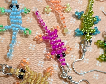 May include: A selection of beaded lizard earrings in shades of blue, orange, green, yellow, and purple. Each earring showcases a lizard design with silver ear wires. The earrings are presented on a peach-coloured fabric.