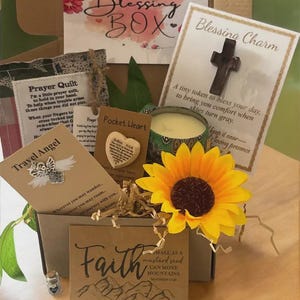 May include: A Blessing Box filled with inspirational items. Includes a candle, a sunflower, a cross charm, a pocket heart, a travel angel, and a card with the word "Faith" and a mountain illustration. The box also contains a "Prayer Quilt" card.