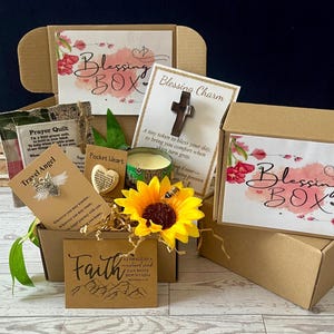 May include: A brown cardboard Blessing Box filled with various items. Includes a cross charm, a prayer quilt, a travel angel pin, a pocket heart, a candle, and a sunflower. The box has the words "Blessing Box" printed on it.