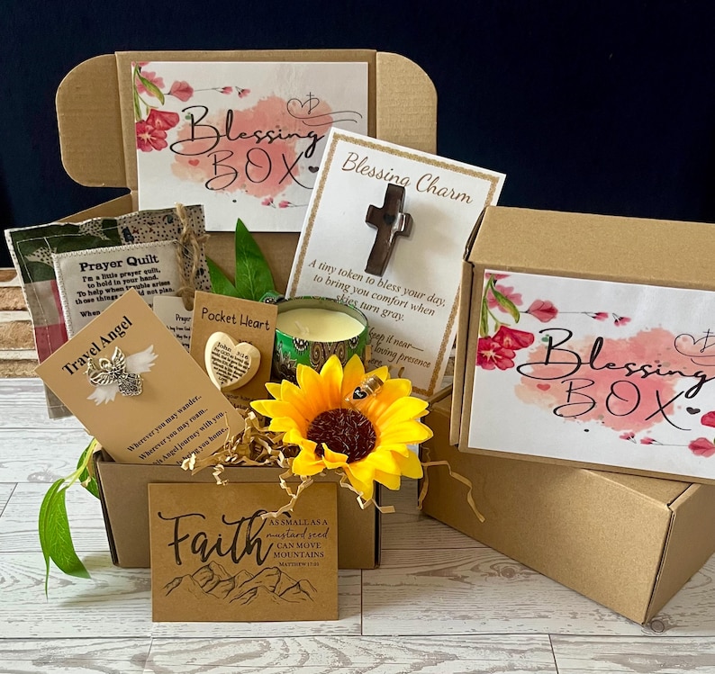 May include: A brown cardboard Blessing Box filled with various items. Includes a cross charm, a prayer quilt, a travel angel pin, a pocket heart, a candle, and a sunflower. The box has the words "Blessing Box" printed on it.