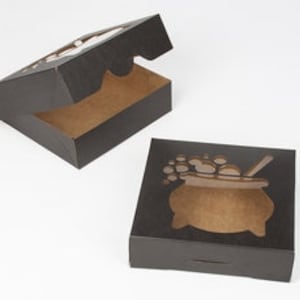 Puede incluir: Caja de cartón negra con un diseño recortado de un caldero con burbujas y una cuchara para remover. La caja está abierta y el interior es marrón.