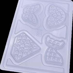 May include: A clear plastic mold with four different shapes: a butterfly, a heart, a diamond, and a mermaid tail. Each shape is designed with circular indentations, likely for creating a textured surface. The text "DMB 3000" is visible.