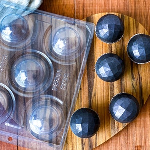 Puede incluir: Un molde de plástico transparente con seis cavidades redondas para hacer caramelos de chocolate. Seis caramelos de chocolate negro están dispuestos en una tabla de madera, cada uno con un diseño facetado.