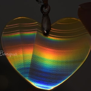 May include: A heart-shaped pendant with a rainbow effect, reflecting colours of yellow, orange, and blue. The pendant is suspended from a silver-coloured bail. The text "© Bonqua" is visible on the pendant.