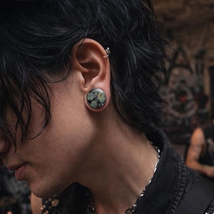 May include: A large, round ear plug with a dark green and black speckled design. The plug is set in a silver-colored metal frame. The image shows the plug in an ear, with dark hair and a silver hoop earring visible.