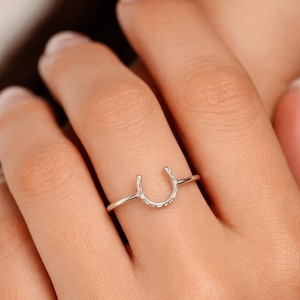 May include: A silver horseshoe ring. The ring features a delicate horseshoe design with a textured surface. The ring is worn on a finger, showcasing its simple and elegant style. The ring is a symbol of good luck and fortune.