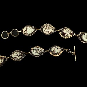 K&ouml;nnte beinhalten: Ein silberfarbenes Armband mit tropfenf&ouml;rmigen Steinen. Jeder Stein hat eine cremefarbene Basis mit brauner Marmorierung. Die Steine sind in einer silbernen L&uuml;nette mit einem Perlenrand gefasst. Das Armband hat einen Knebelverschluss und einen Ringverschluss.
