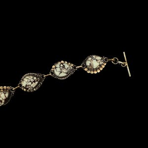 K&ouml;nnte beinhalten: Ein silbernes Armband mit tropfenf&ouml;rmigen Steinen. Die Steine sind wei&szlig; mit dunkelbraunen Adern, eingefasst in Silber mit einem goldfarbenen, perlenbesetzten Rand. Das Armband hat einen Knebelverschluss.