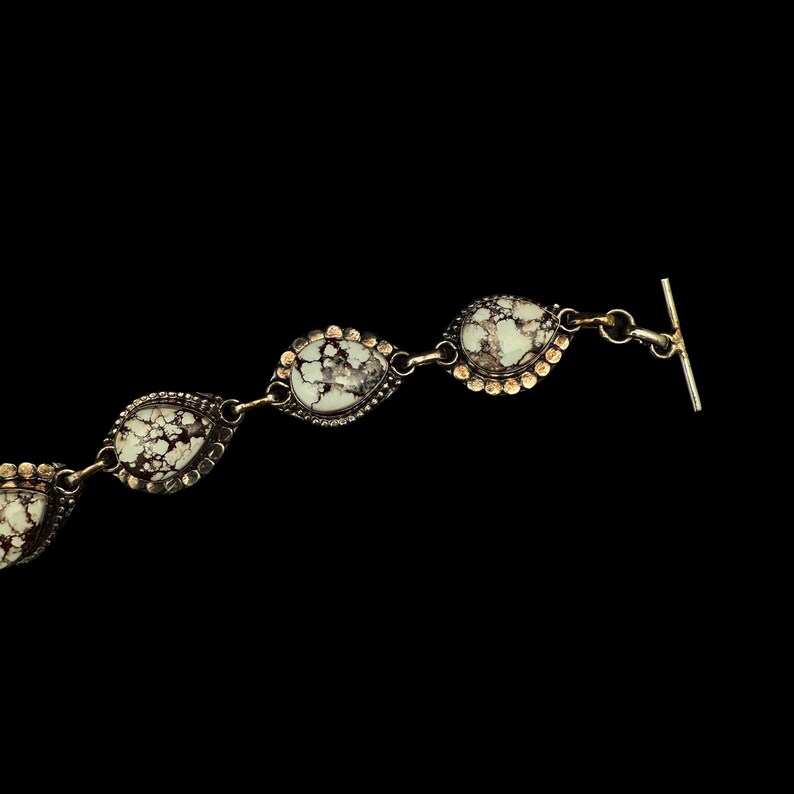 K&ouml;nnte beinhalten: Ein silbernes Armband mit tropfenf&ouml;rmigen Steinen. Die Steine sind wei&szlig; mit dunkelbraunen Adern, eingefasst in Silber mit einem goldfarbenen, perlenbesetzten Rand. Das Armband hat einen Knebelverschluss.