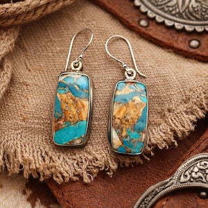 May include: A pair of silver-toned earrings with rectangular turquoise and gold-colored stone inlays. The earrings have a hook closure and are displayed on a textured brown surface, showcasing the jewelry's natural stone patterns and metallic accents.