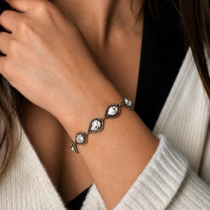 K&ouml;nnte beinhalten: Ein goldfarbenes Armband mit tropfenf&ouml;rmigen wei&szlig;en Steineinfassungen mit schwarzen Adern. Das Armband wird am Handgelenk getragen, mit einem cremefarbenen Pullover und einem schwarzen Oberteil im Hintergrund. Das Armband hat eine Vintage- oder Antik-&Auml;sthetik.
