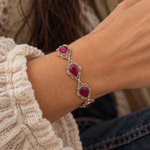 Puede incluir: Una pulsera de eslabones de plata con varias piedras preciosas de color rosa intenso en forma de lágrima. Cada piedra está engastada en un marco de plata intrincado y ornamentado con detalles metálicos, unidos por pequeñas anillas de plata.