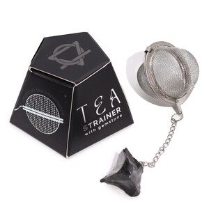May include: A black pyramid-shaped box with the words "TEA STRAINER with gemstone" and a silver tea infuser with a chain and a black gemstone. The box has a logo on top and the words "mistical tea experience".