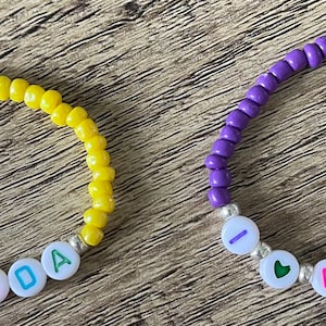 May include: Two beaded bracelets. One is yellow with the word "DADA" spelled out in colorful letters. The other is purple with the word "MAMA" spelled out in colorful letters. Both bracelets have the words "I ❤️" on them.