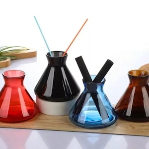 May include: Four glass diffuser bottles in different colours: red, black, blue, and brown. The bottles are cone-shaped and have a wide opening at the top. The bottles are arranged in a row on a wooden surface.