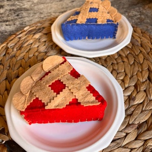 May include: Two felt pie slices on white plates. One pie slice is red with a brown crust and the other is blue with a brown crust.