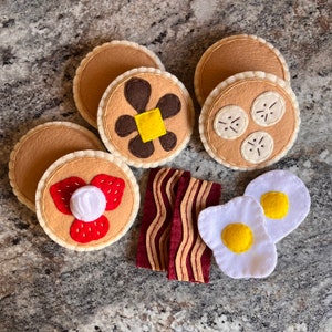 May include: Felt play food set with a variety of breakfast items, including pancakes, bacon, and eggs. The pancakes are decorated with strawberries and chocolate chips. The bacon is brown and crispy. The eggs are white and yellow.
